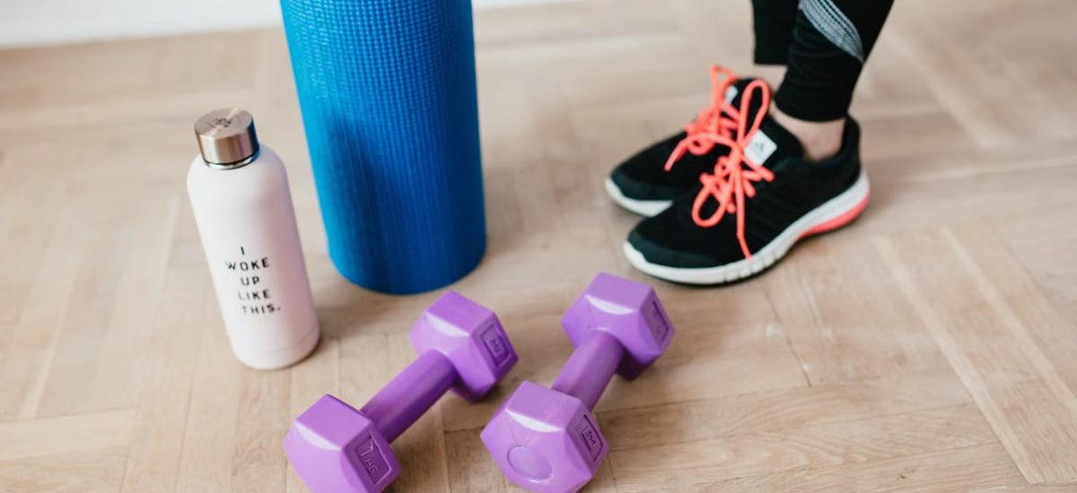 Unrecognizable sportswoman wearing sneakers and black leggings standing with sport mat on wooden floor near dumbbells and water bottle before exercising indoors