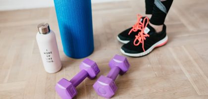 Unrecognizable sportswoman wearing sneakers and black leggings standing with sport mat on wooden floor near dumbbells and water bottle before exercising indoors