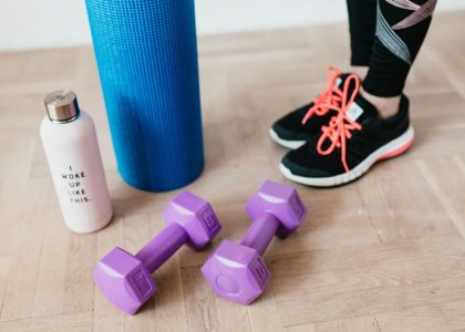 Unrecognizable sportswoman wearing sneakers and black leggings standing with sport mat on wooden floor near dumbbells and water bottle before exercising indoors